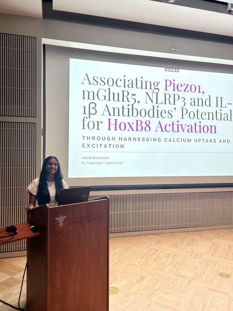A woman stands at a podium in front of a projection screen displaying a scientific presentation titled 'Associating Piezo1, mGluR5, NLRP3 and IL-1β Antibodies' Potential for HoxB8 Activation. The podium has a symbol and text indicating it is from the University of Illinois College of Medicine.