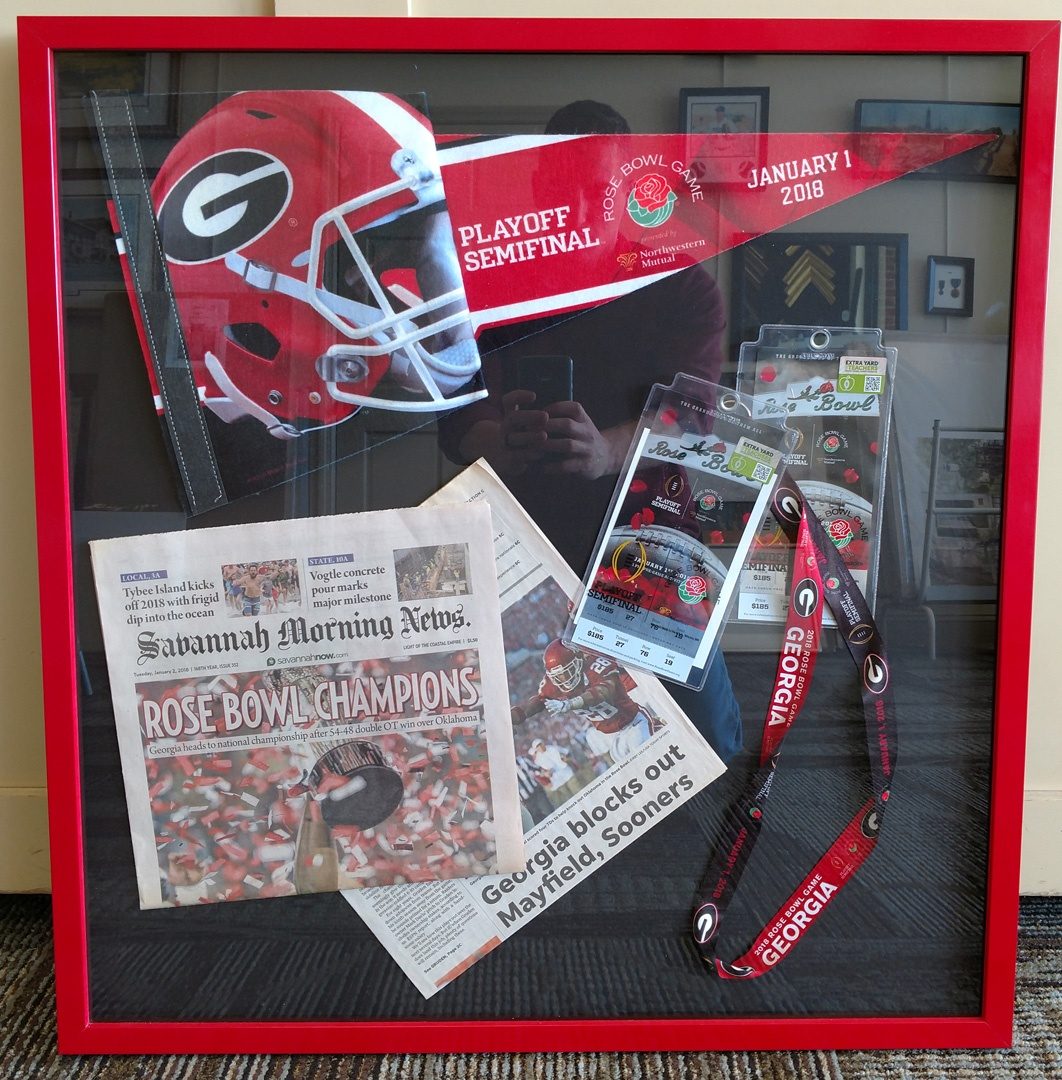 A red-framed shadow box holding a Georgia Bulldogs Rose Bowl pennant, game tickets, a lanyard and the Savannah Morning News front page reading Rose Bowl Champions