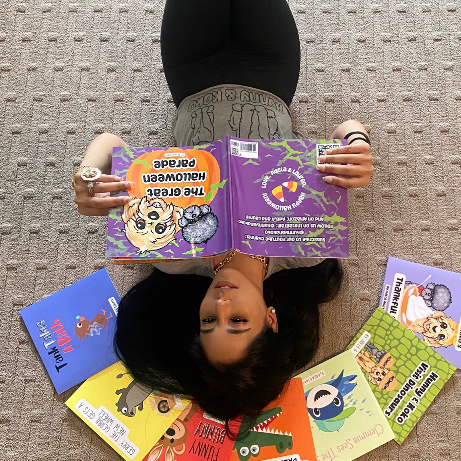 A woman lying on the carpeted floor, holding an illustrated children's book titled "The Great Halloween Parade" upside down. She is surrounded by colorful children's books with cartoon characters on the covers, including a book with a rat and another with a frog. The woman has dark hair, is wearing a black top, beige shorts, and jewelry including a ring and a chain necklace.