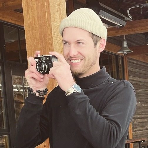 Adrian Hallberg in a knit beanie, smiling while shooting with a compact film camera
