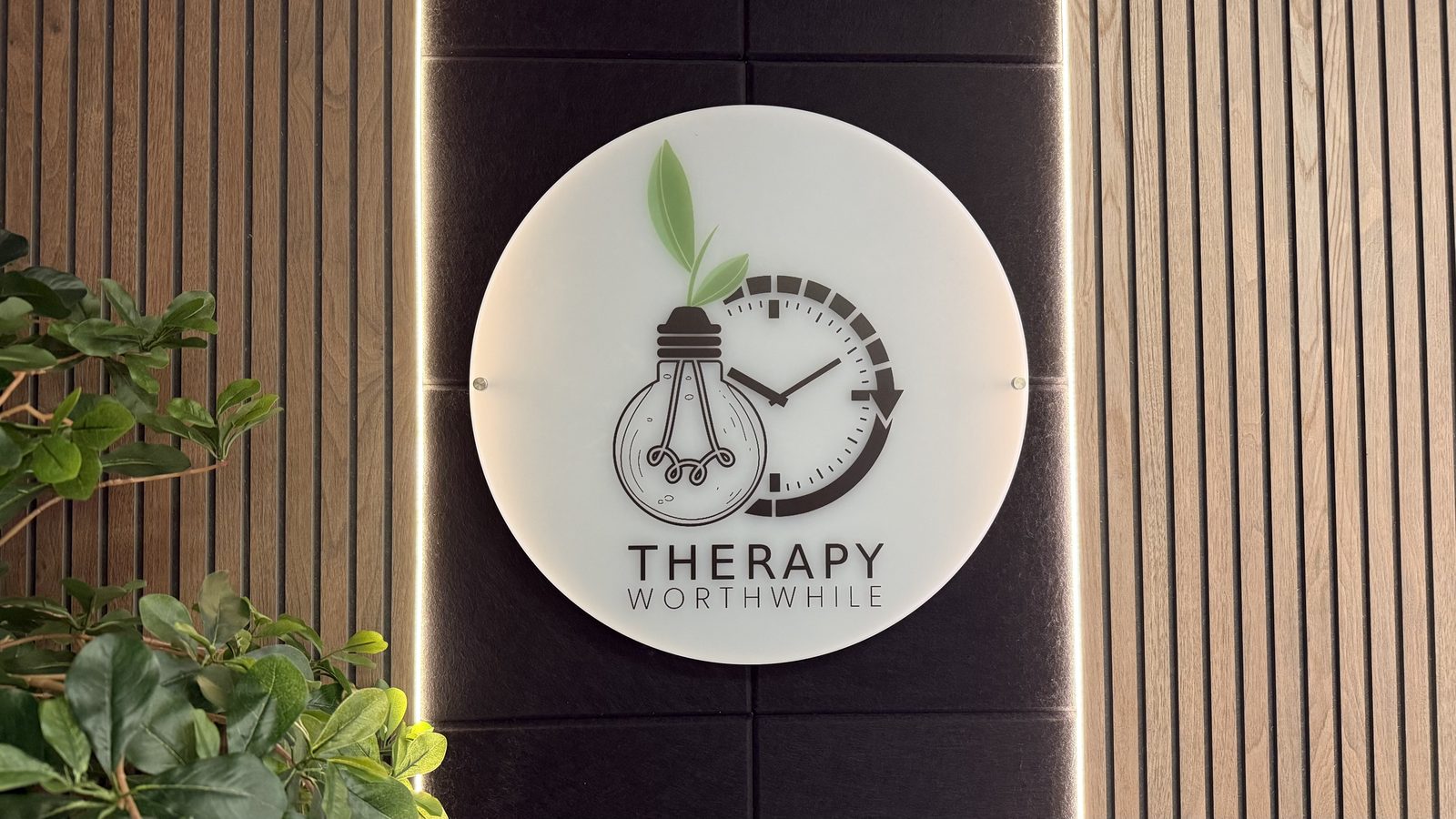 Round Therapy Worthwhile sign on a slatted wood wall, a light bulb sprouting a leaf beside a clock