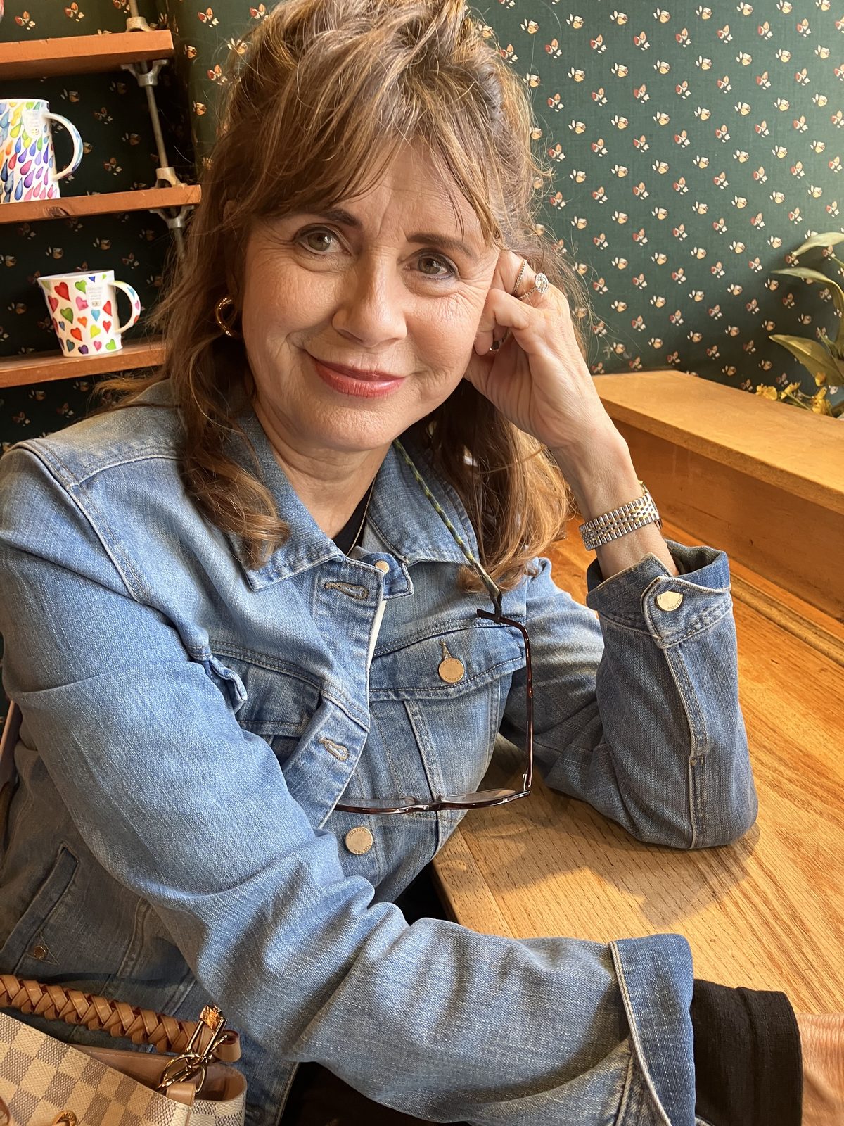 Amy Mink, founder of Amy Mink Design, smiling in a denim jacket at a wooden table in front of green floral wallpaper