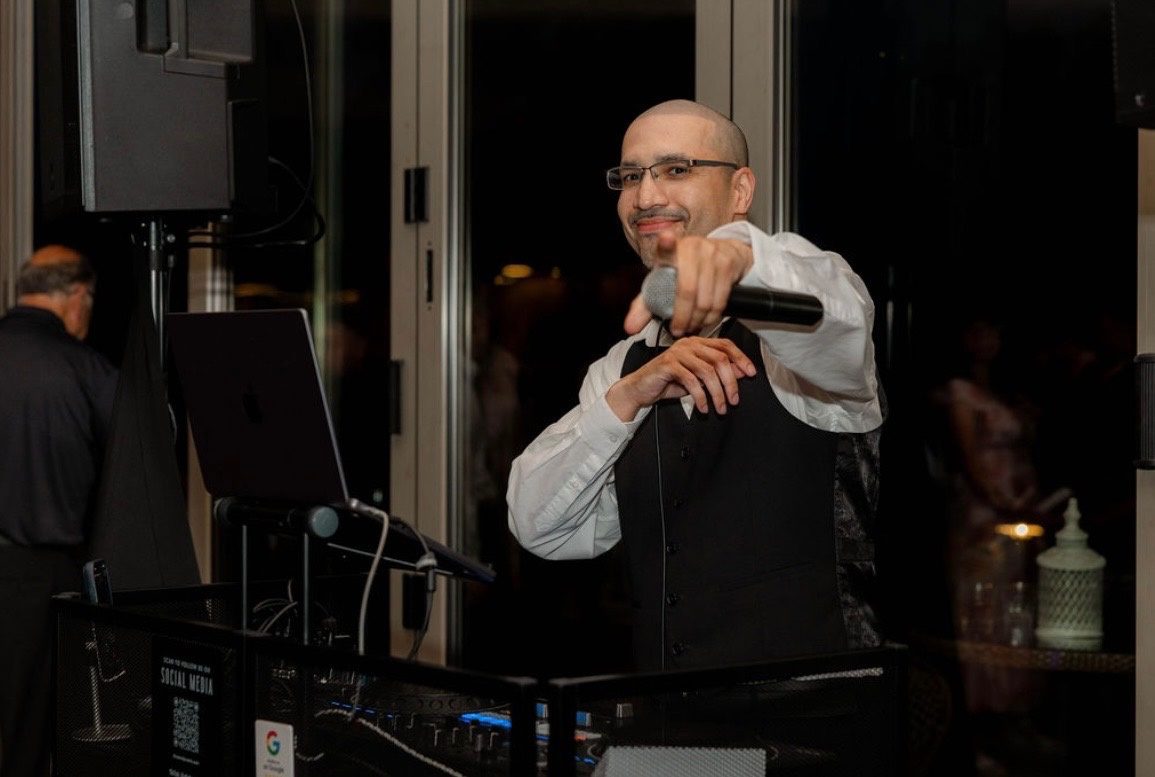 Angel Rivera behind the DJ booth at an event, pointing a microphone toward the camera