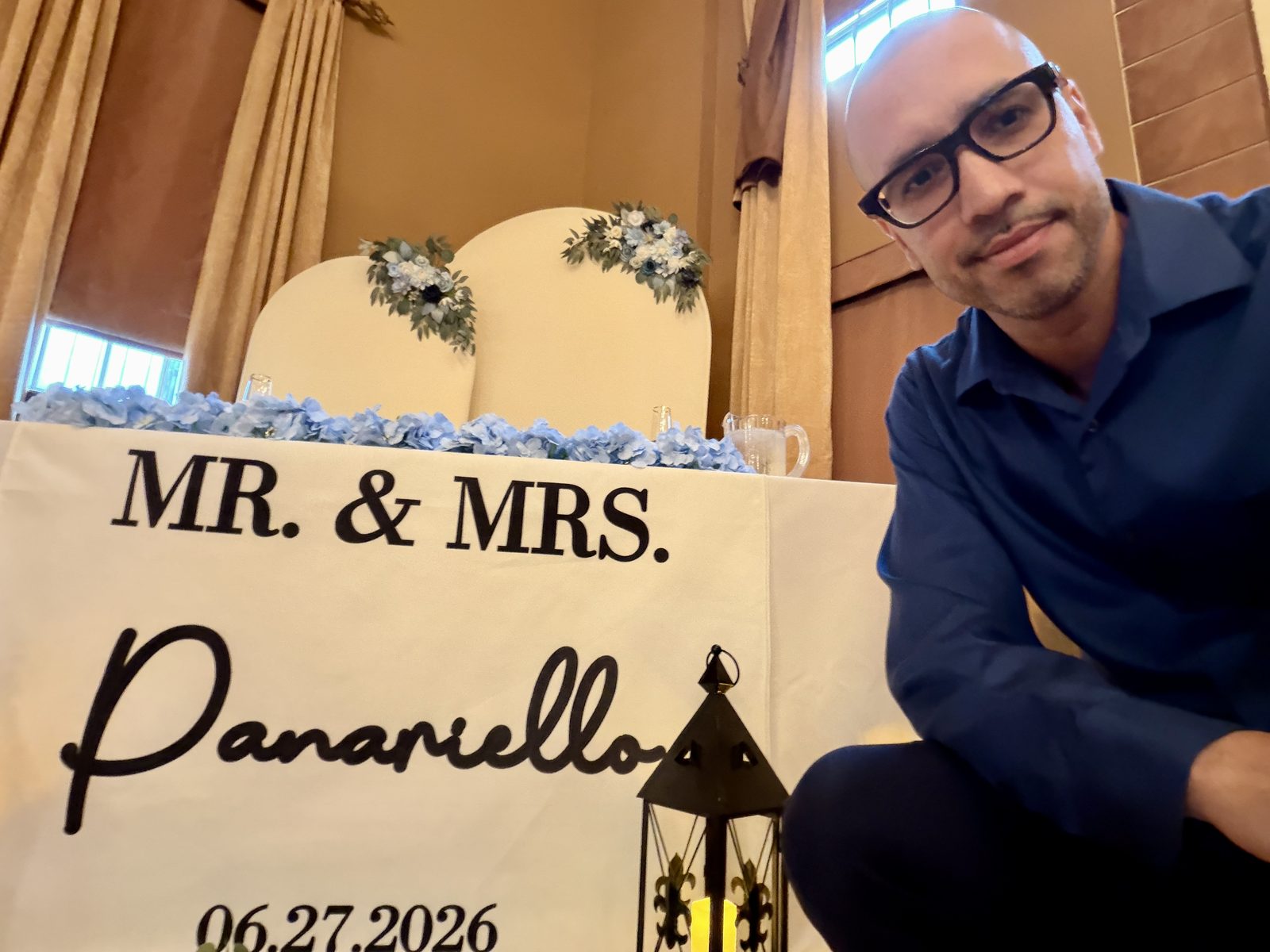 Angel Rivera beside a sweetheart table decorated with blue hydrangeas and a Mr. & Mrs. Panariello sign at a wedding reception
