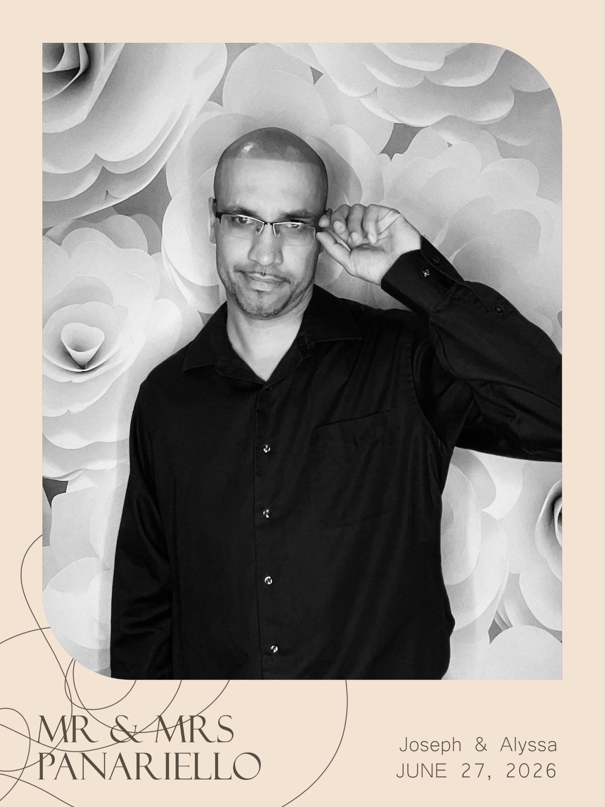 Black and white photo booth print of Angel Rivera adjusting his glasses in front of a paper flower wall at a wedding