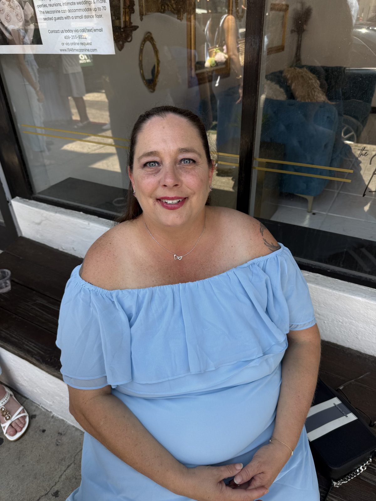Angie Stewart, owner of Dallas Placenta Encapsulation, smiling in a light blue off-the-shoulder dress outside a storefront in Sanger, Texas