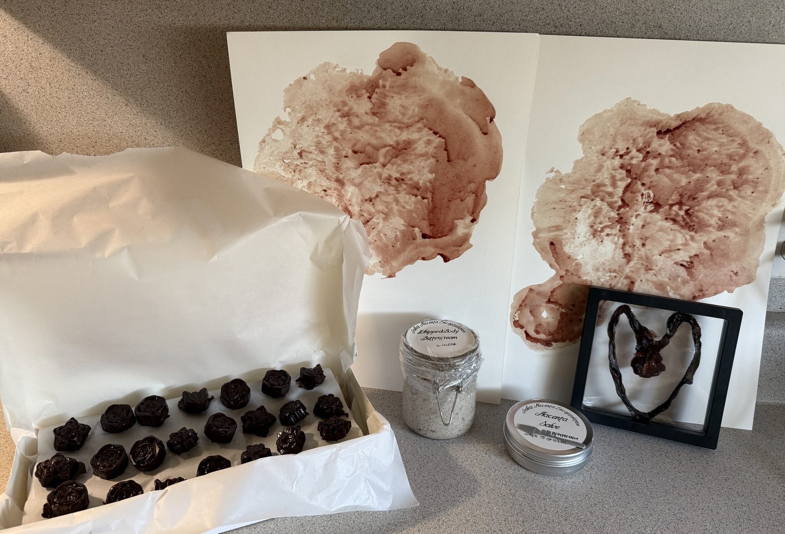 A box of placenta-infused chocolates beside two placenta prints, a jar of capsules, a tin of salve and a framed heart-shaped keepsake