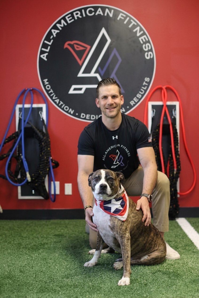 Antonio Somma Jr., founder of All-American Fitness in Morris Plains, New Jersey, kneeling with a dog on the gym's turf