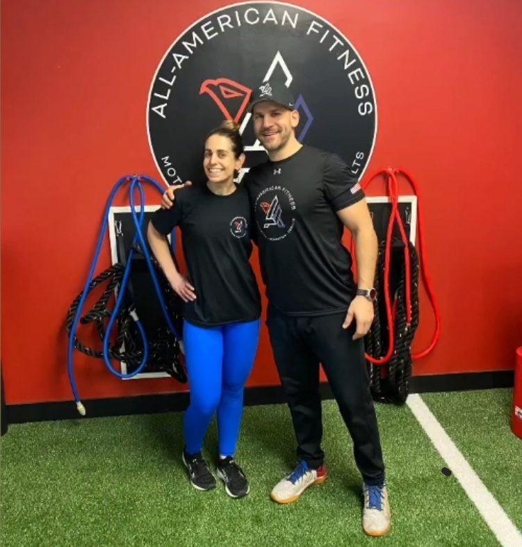 Antonio Somma Jr. with a member of the All-American Fitness community in front of the gym's logo wall