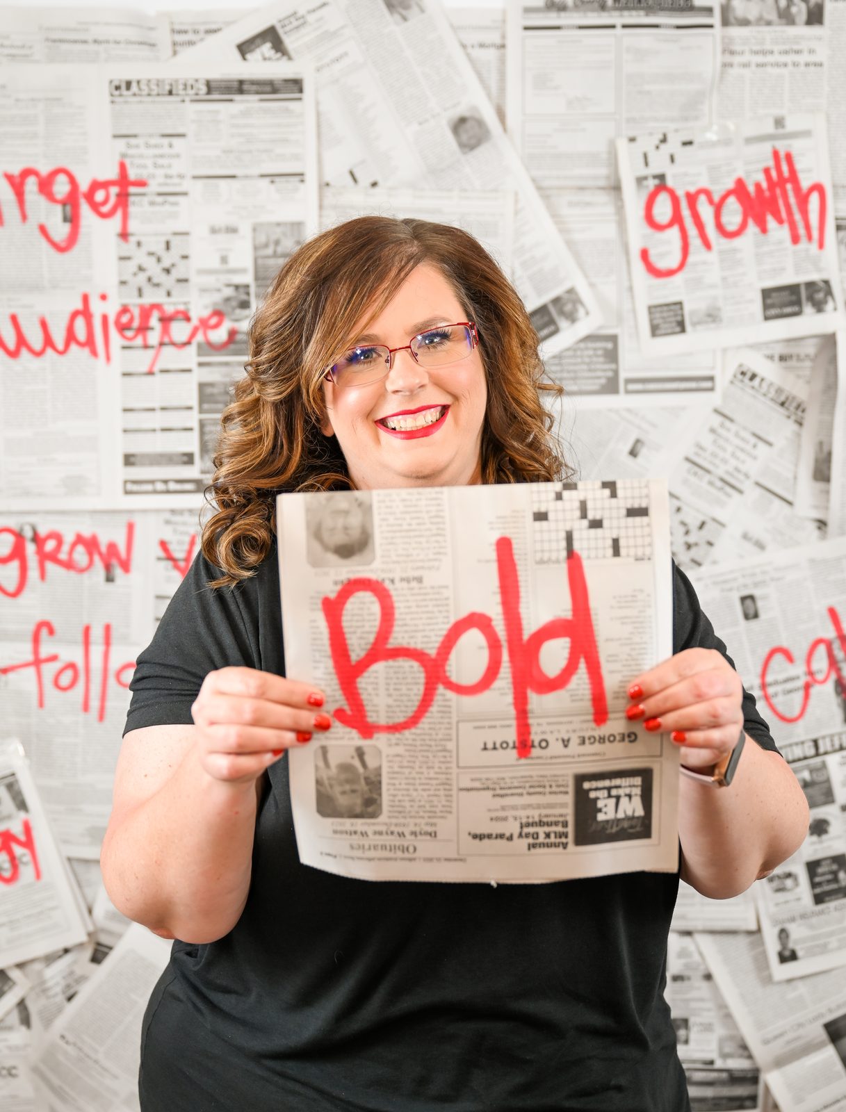 April Barnard, founder of Marketing with April, smiling and holding up a newspaper page marked "Bold" in red, in front of a wall of newspapers