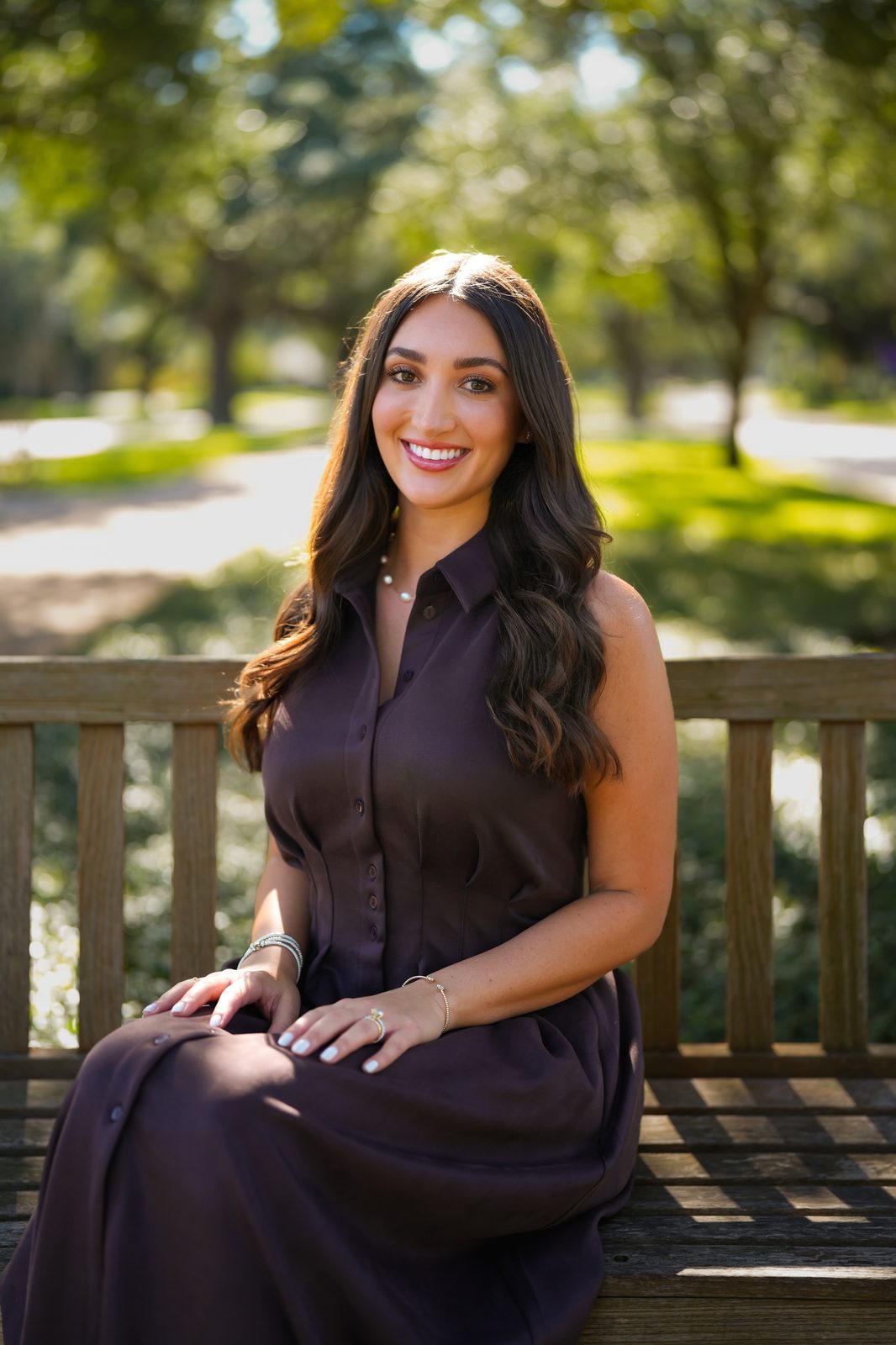 Ariana Stanley seated on a wooden park bench in a dark brown dress on a sunny day in Houston