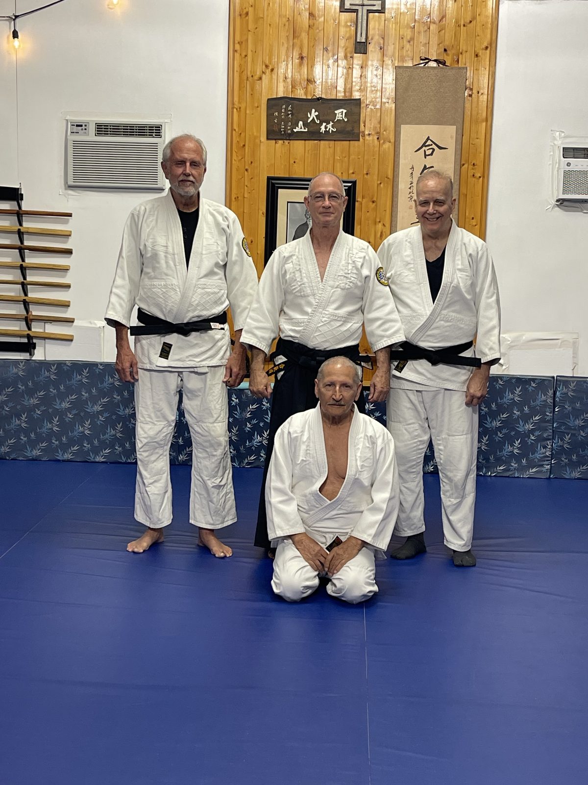 Bill Hague standing with two fellow black belts behind a kneeling senior teacher on the dojo mat
