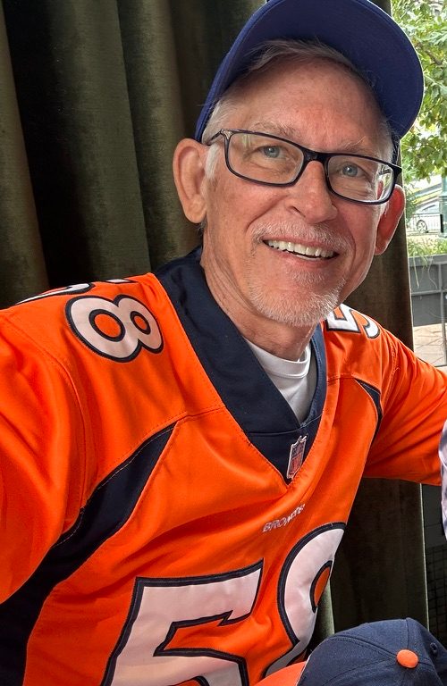 Bret Miller, founder of Sports History Group, smiling in glasses, a blue cap and an orange Denver Broncos jersey