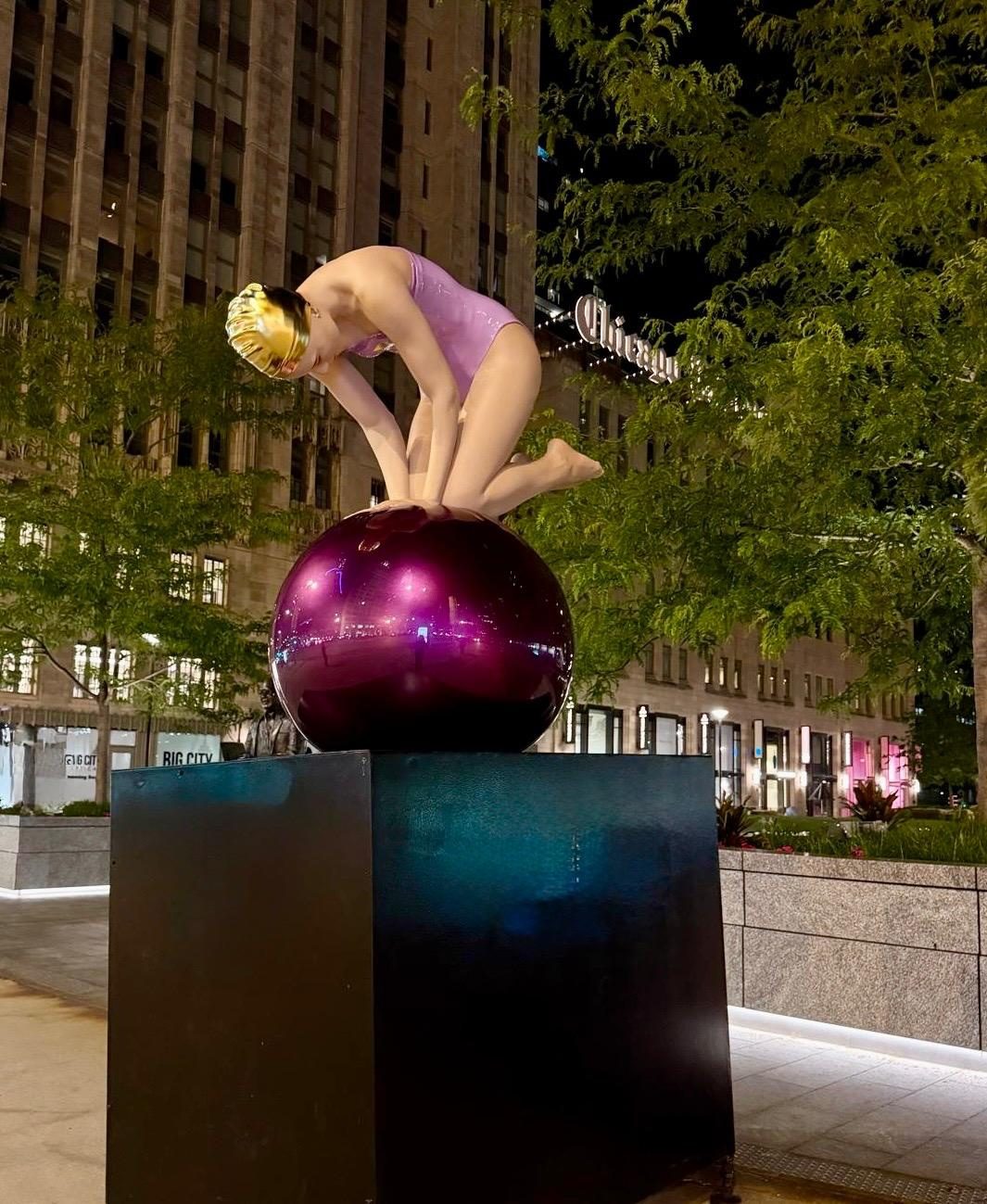 Quan, a Carole Feuerman sculpture of a swimmer in a gold cap crouched on a purple sphere, lit at night in Chicago