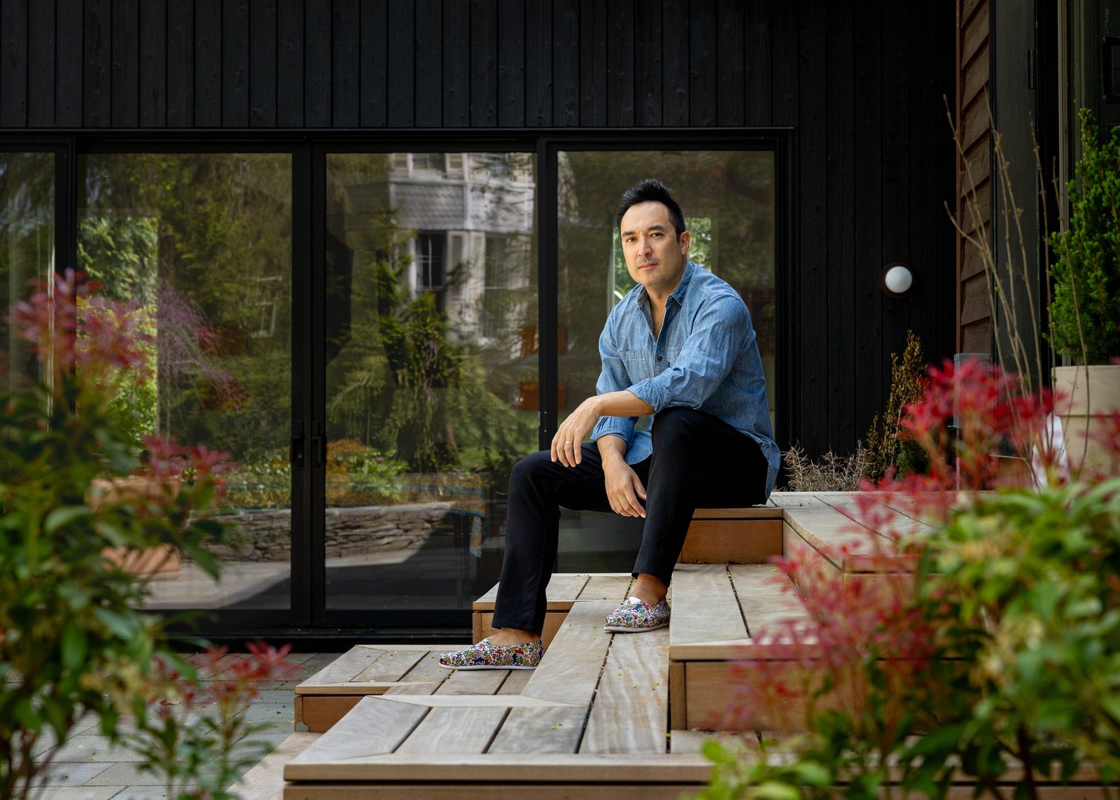 Chris Kao sitting on the stepped timber deck of a black-clad house, surrounded by red-tipped shrubs