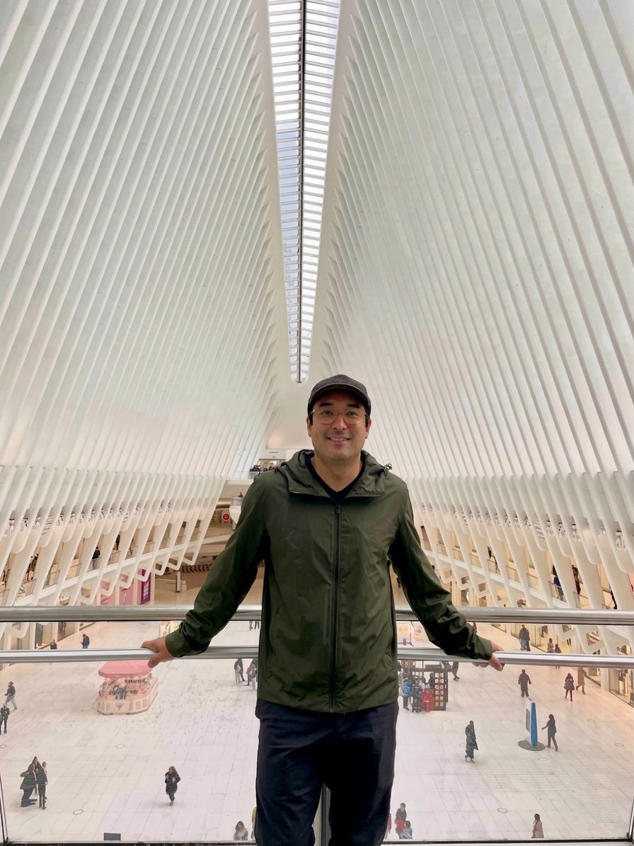 Chris Kao in a green jacket on a balcony inside the Oculus at the World Trade Center, white ribs rising overhead