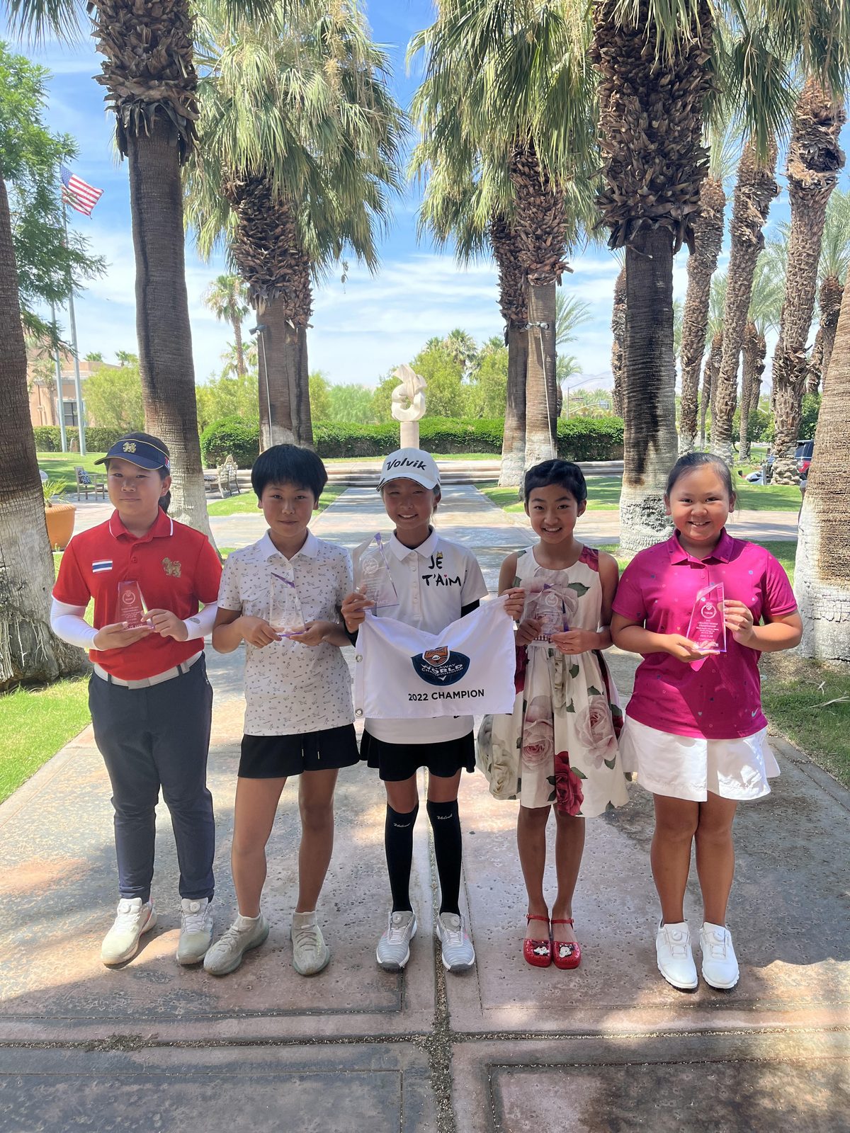 Five junior golfers holding trophies and an FCG champion flag beneath palm trees