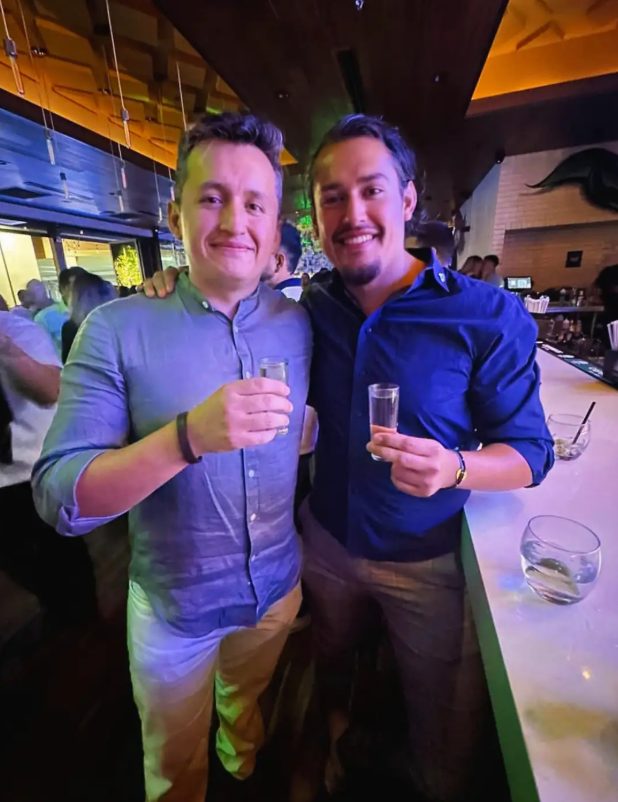 Two men smiling and holding shot glasses in a lively bar or nightclub with colorful lighting. One has curly hair and the other has straight dark hair. Background shows other people and a bar area.