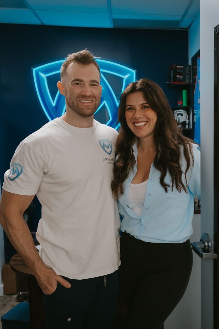Crystal Preyer and her husband Stephen Preyer, founders of Armored Massage, smiling in front of the glowing blue Armored Massage logo