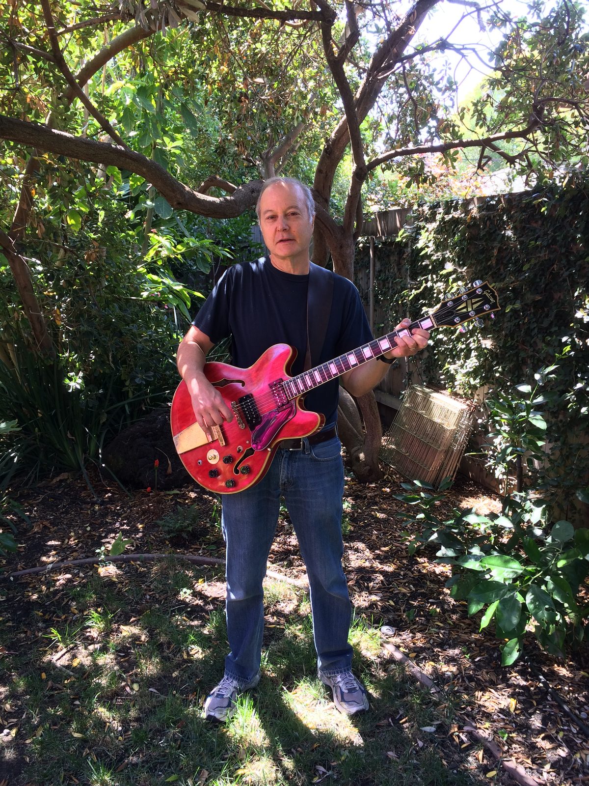Dan Zalles standing in a sunlit garden playing a red Gibson electric guitar