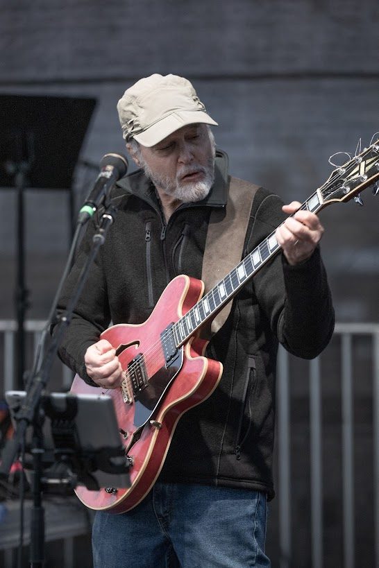 Dan Zalles playing guitar and singing at a microphone on an outdoor stage