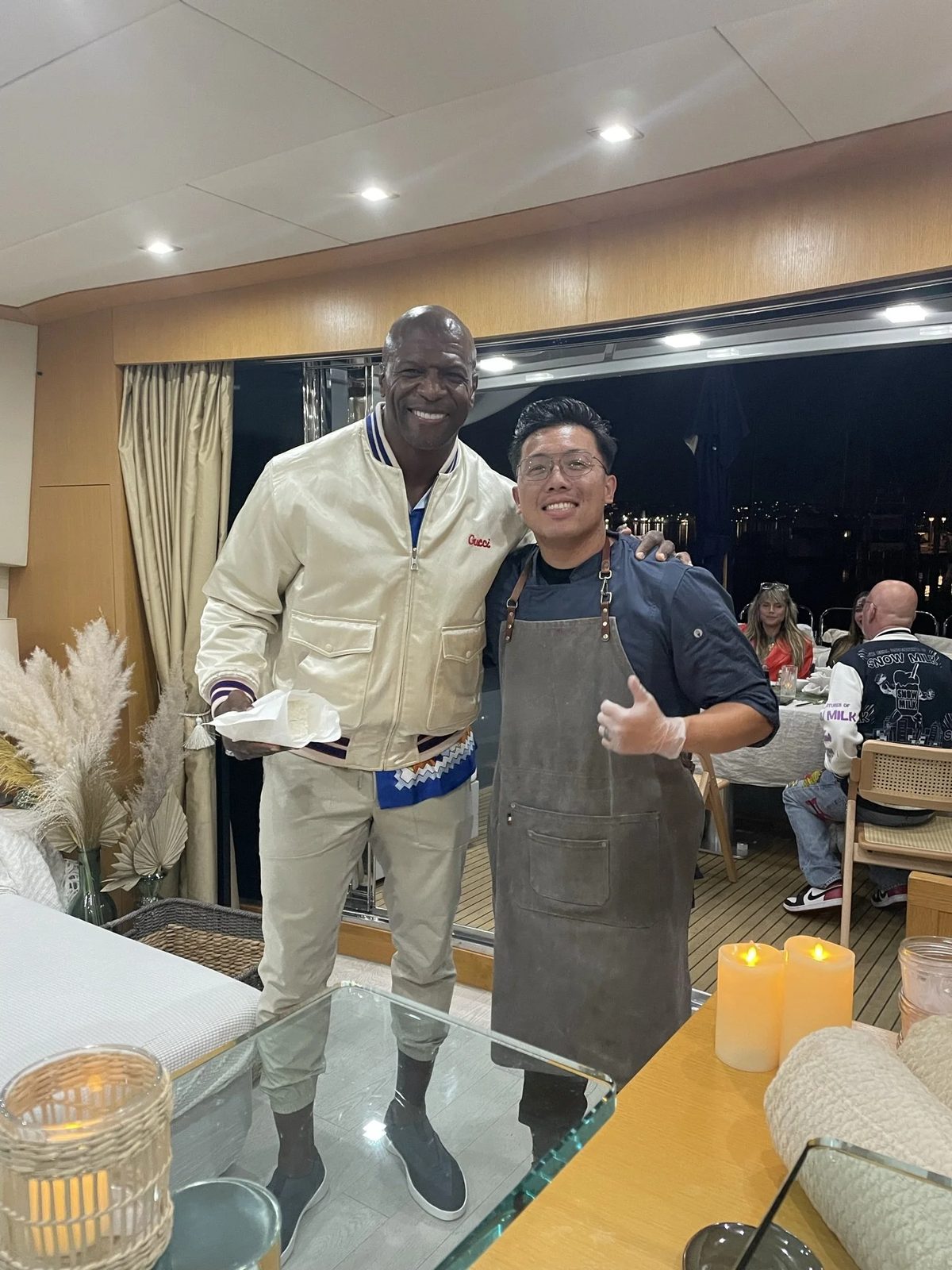 Two men standing and smiling indoors at night, one taller with dark skin and wearing a cream-colored jacket, the other shorter with light skin, glasses, and wearing a dark shirt with an apron. People are dining at tables in the background.