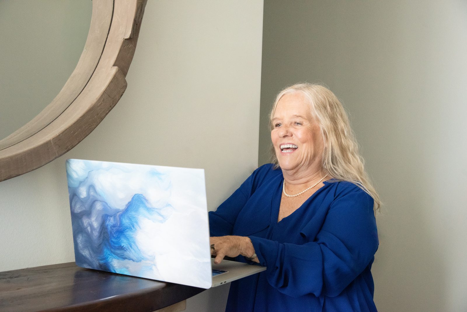 Dianne Allen smiling as she works at a laptop beside a round wooden mirror