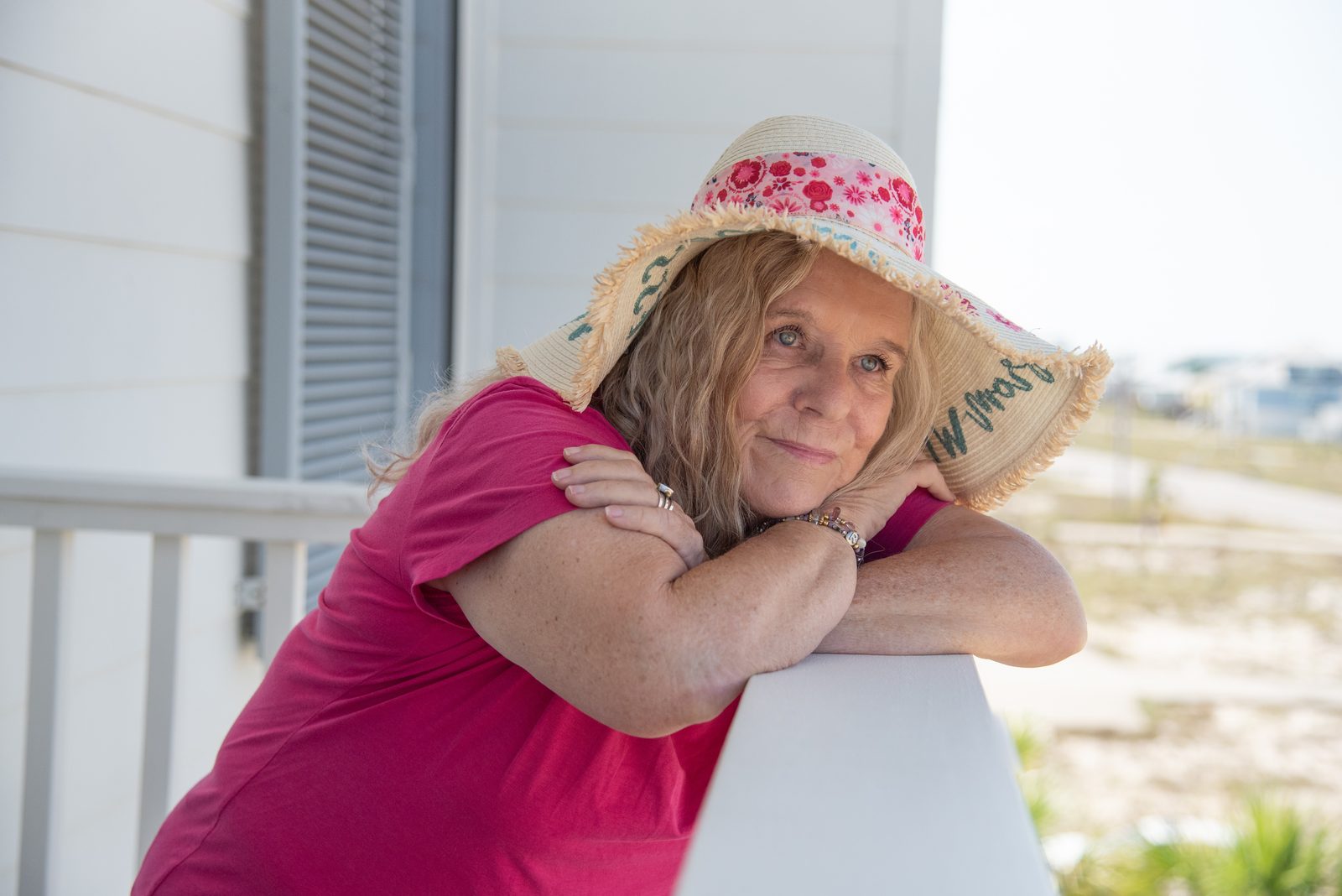 Dianne Allen in a pink shirt and sun hat leaning on a porch railing near the beach