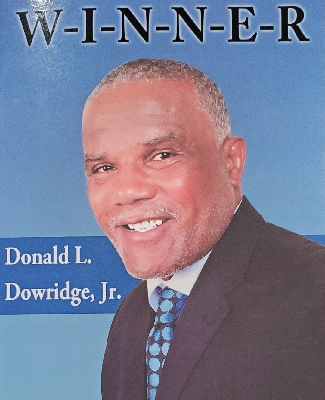 A W-I-N-N-E-R poster with a smiling portrait of Donald L. Dowridge, Jr. in a dark suit and blue dotted tie