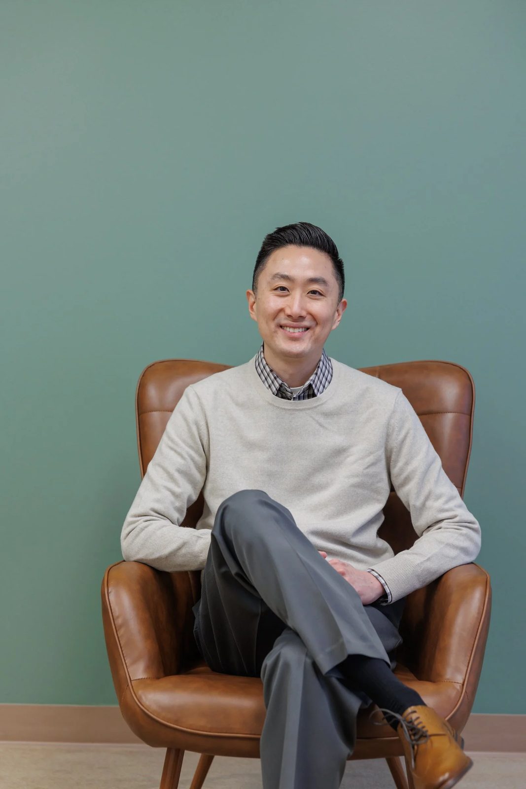 A smiling man sitting in a brown leather chair against a green wall, wearing a beige sweater, checkered shirt, gray pants, black socks, and brown shoes.