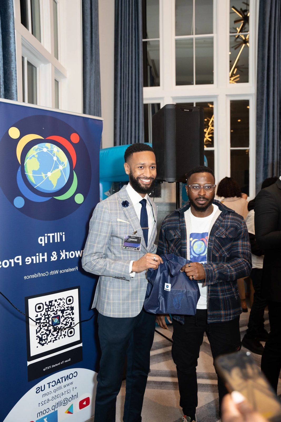 Two men stand together at a conference, one with a gray plaid blazer and the other in a hoodie, holding an event goodie bag. Behind them is a large vertical banner with a globe and network graphics, QR code, and event information.