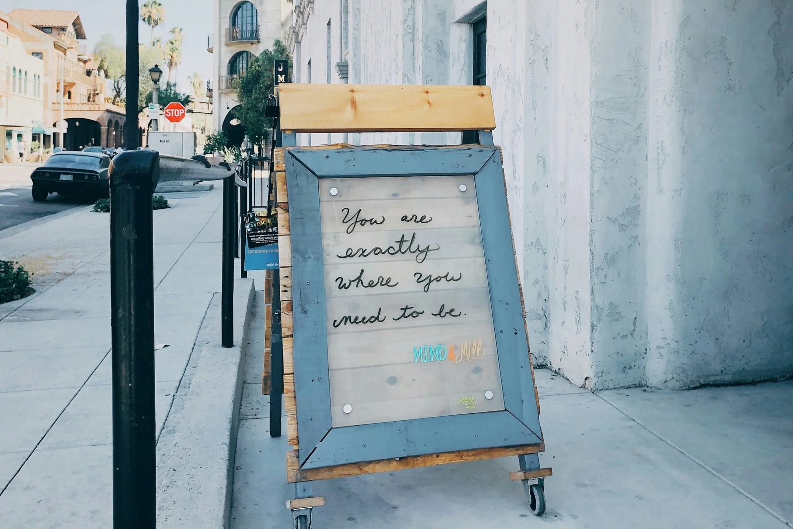 A sidewalk signboard with a quote that says, "You are exactly where you need to be." The sign is placed on wheels and is located next to a concrete wall, with buildings and parked cars visible in the background.