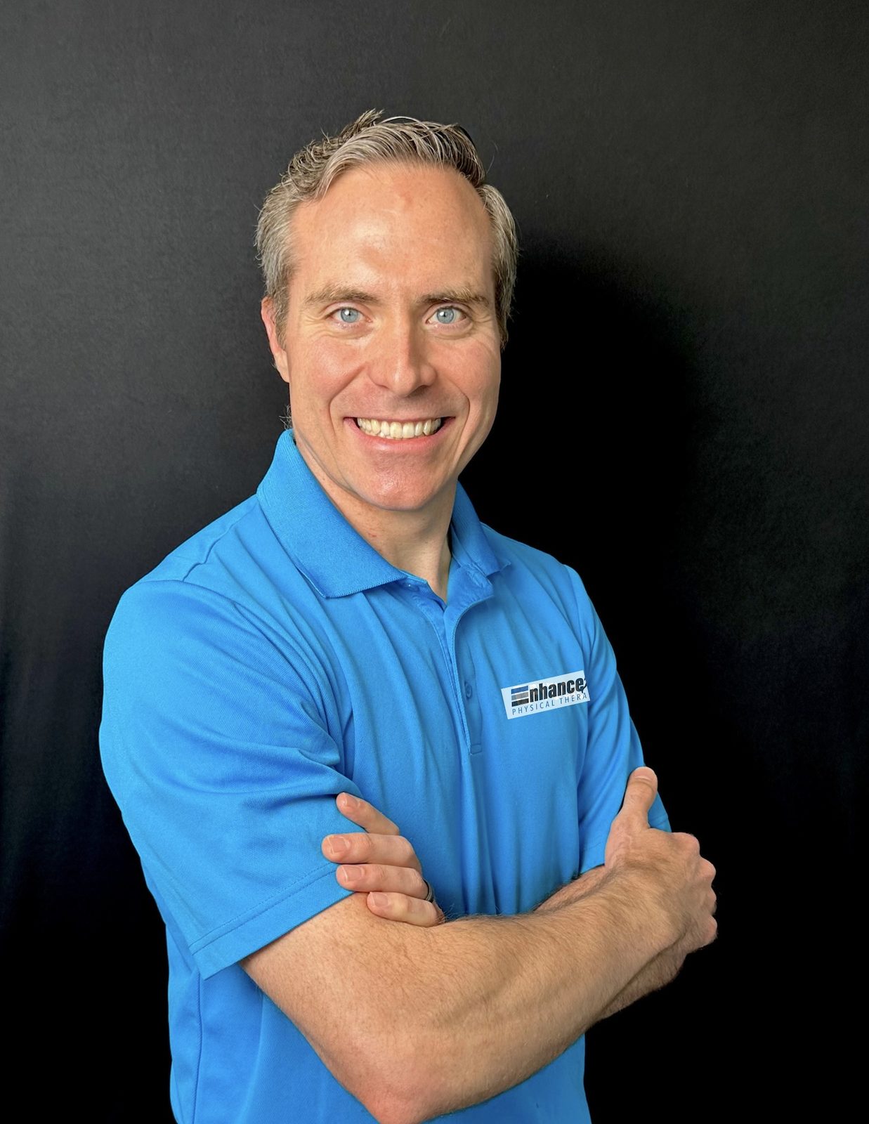 Dr. Doug Ashton, owner of Enhance Physical Therapy, smiling with arms crossed in a blue Enhance polo