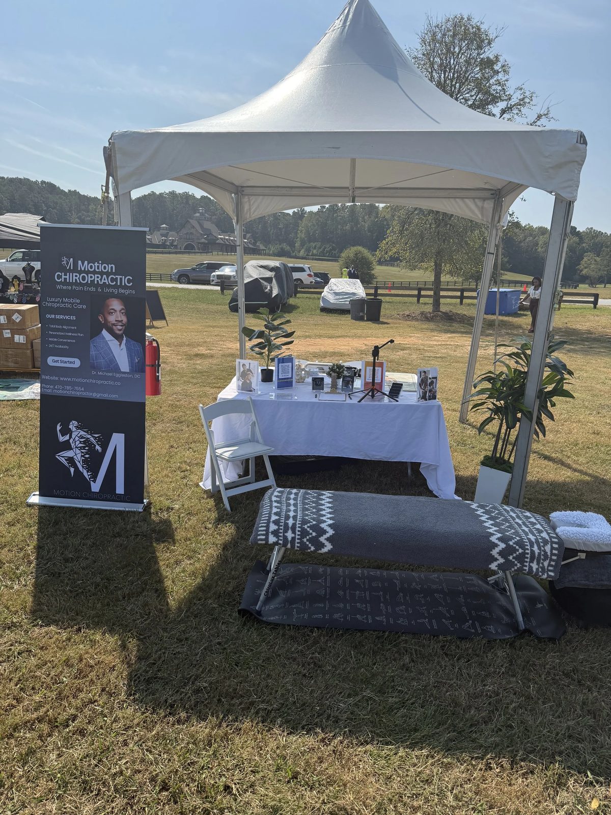 An outdoor booth for Motion Chiropractic under a white canopy tent. The booth has a table with promotional materials, framed photos, and potted plants. A signboard displays information about the chiropractic practice, including a photo of Dr. Michael Eggleston, DC. There is a treatment table with a blanket and towels in front of the booth on the grass.