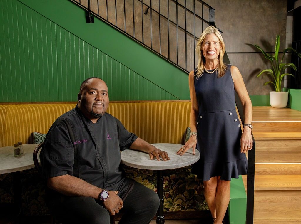 Emily Fournier standing beside Executive Chef Jimmy Norris, seated in chef whites at a marble cafe table under a green staircase