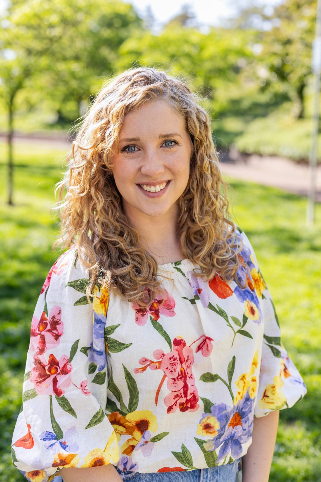 Emma Carpenter, owner of Better Connections Therapy, smiling outdoors in a floral blouse with long curly hair, green trees behind her