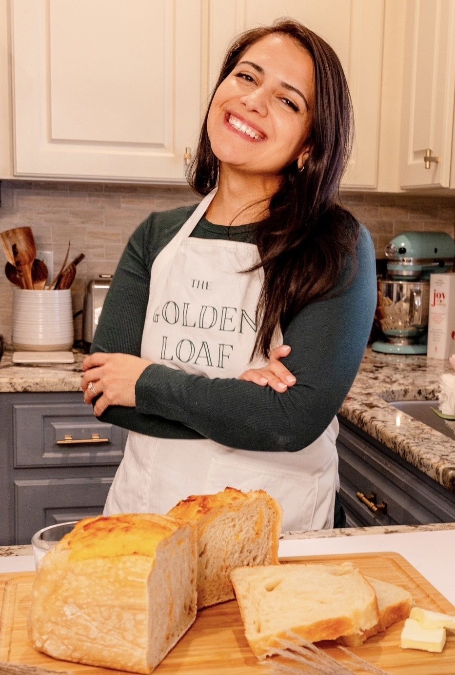 Gaby Miller, owner and baker behind The Golden Loaf Home Bakery, smiling with her arms crossed in her kitchen behind a sliced loaf of sourdough