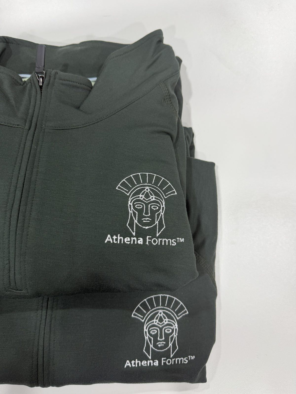 Two folded dark green quarter-zip jackets embroidered with the Athena Forms helmet logo