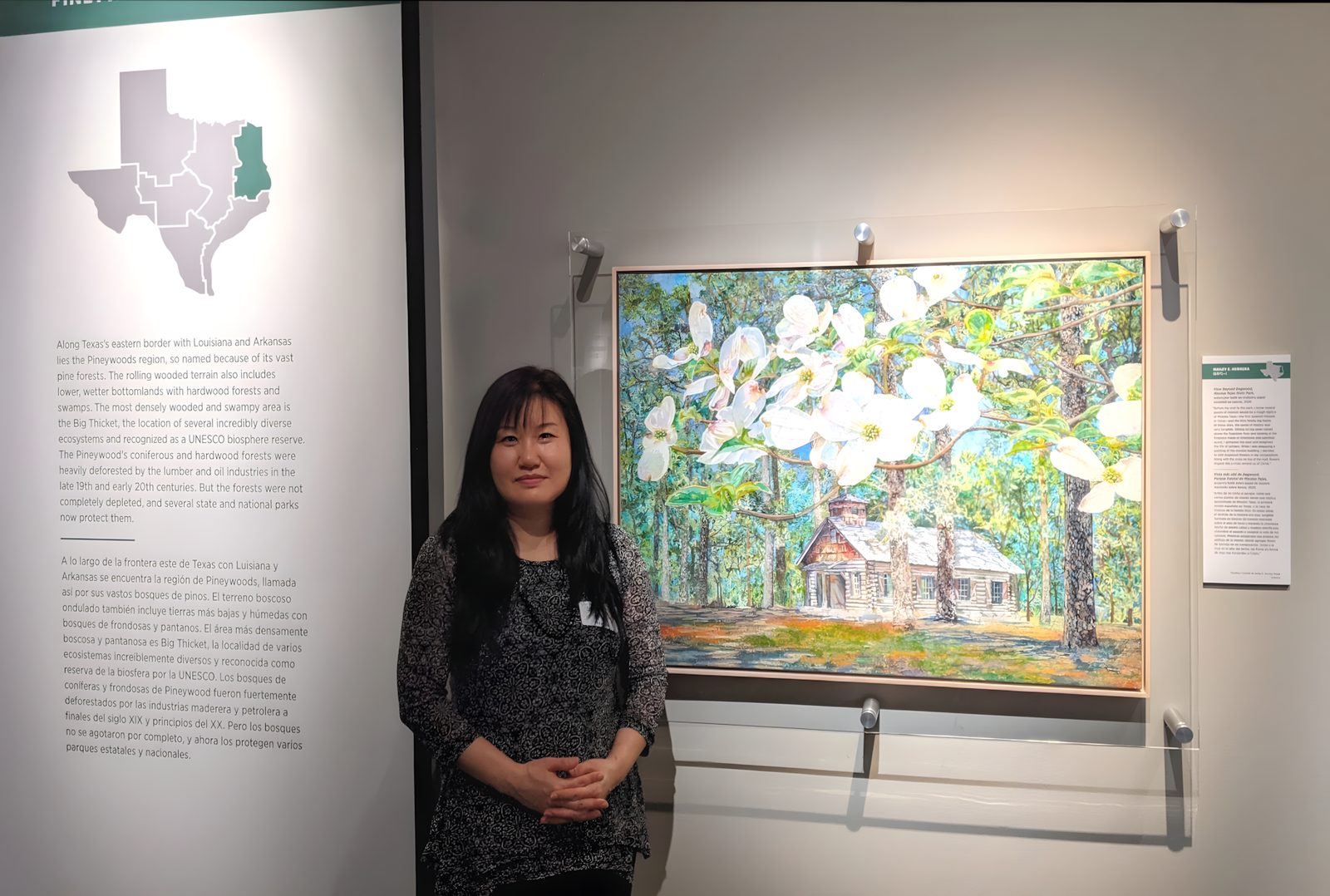 Hailey Herrera standing beside her large watercolor batik painting of white dogwood blossoms and a log cabin in a gallery exhibit about the Texas Piney Woods