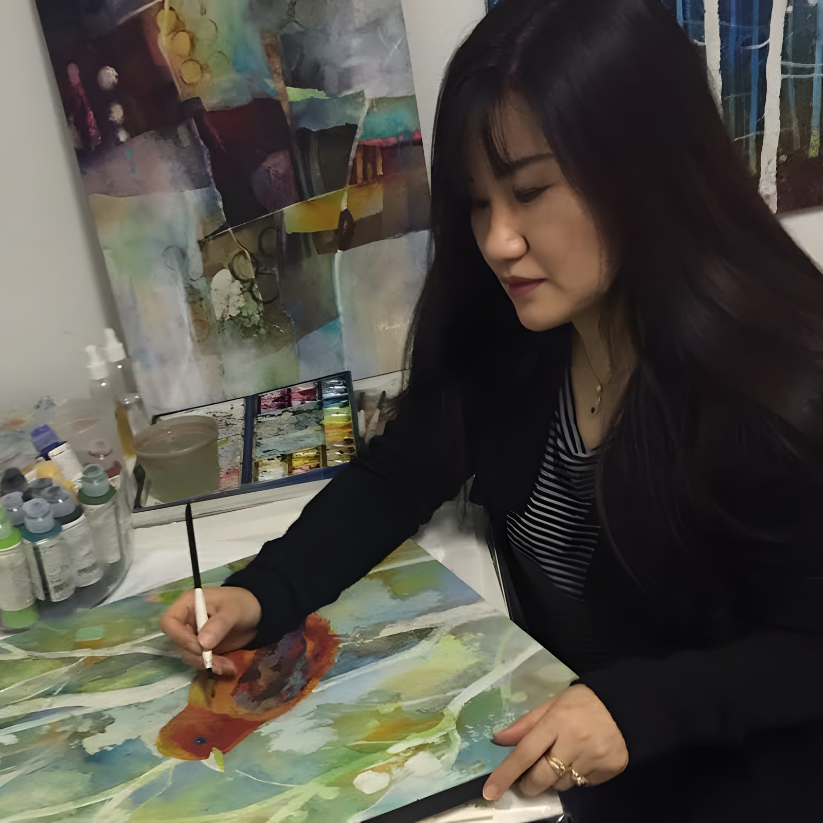 Hailey Herrera painting a red bird onto a green watercolor batik piece in her studio, with paint bottles and a palette beside her
