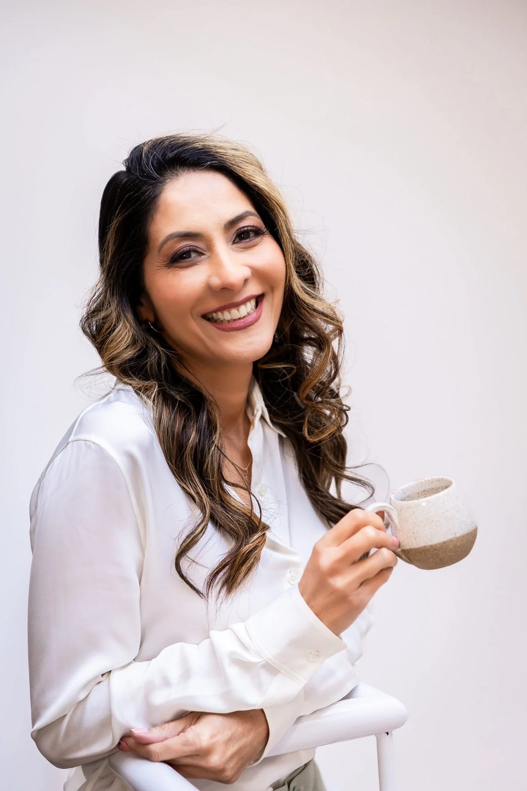 A woman with long wavy brown hair, smiling and holding a ceramic mug, wearing a white blouse, against a plain light background.