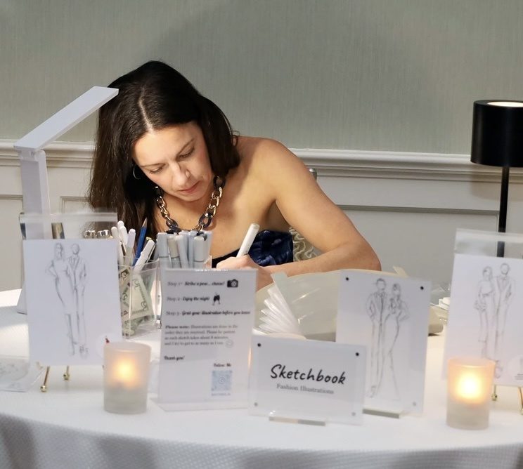 Kaitlin Mahony live sketching guests at her Sketchbook Fashion Illustrations table during an event, with finished sketches on easels and candles