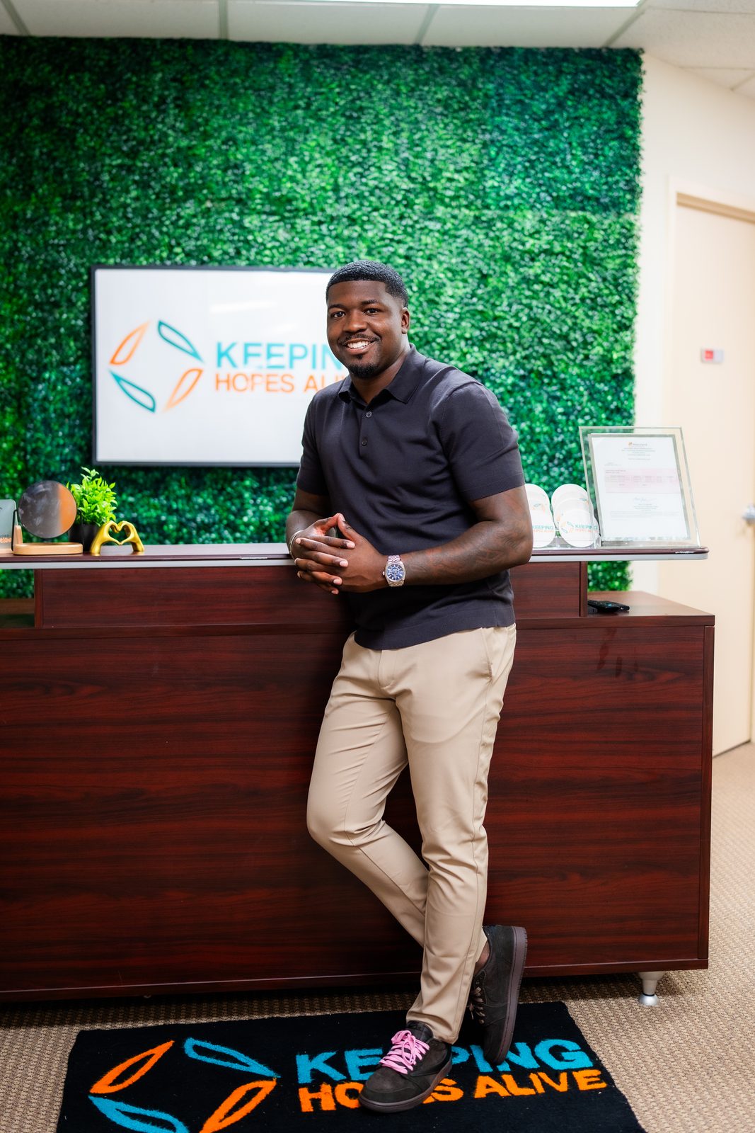 Karahn Alston leaning on the front desk at Keeping Hopes Alive in Baltimore, in front of a green living wall with the clinic's logo