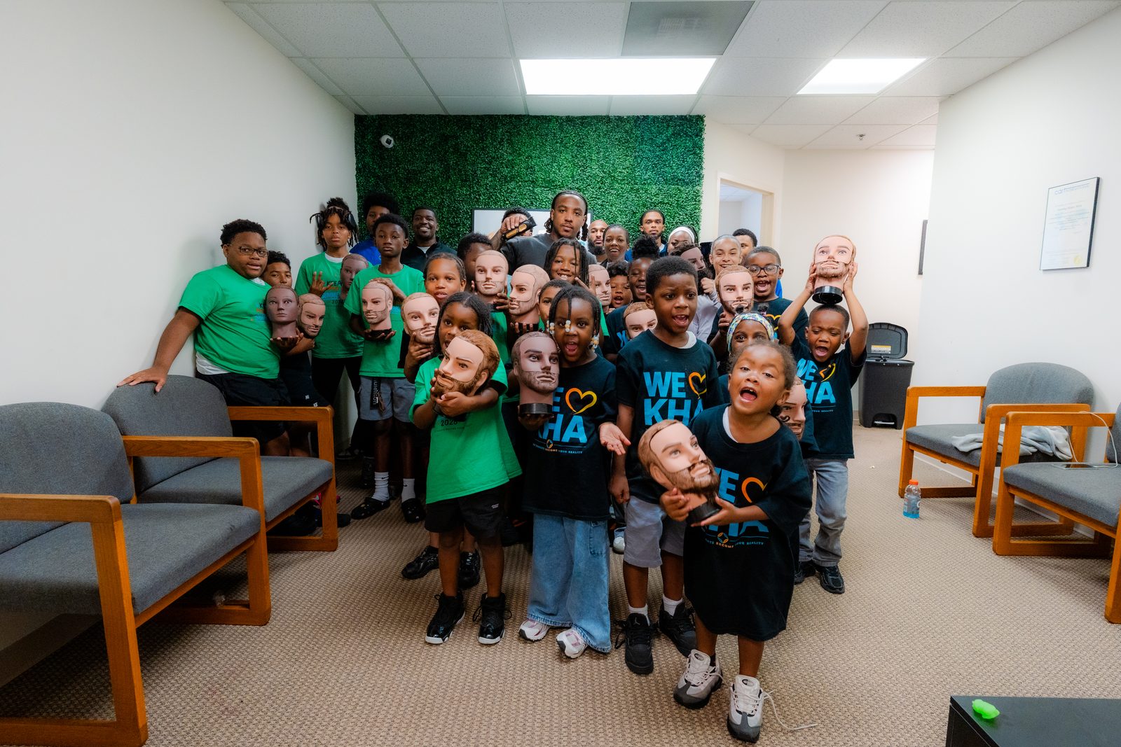 A crowd of children in green and black We Love KHA shirts holding mannequin heads at the Keeping Hopes Alive office
