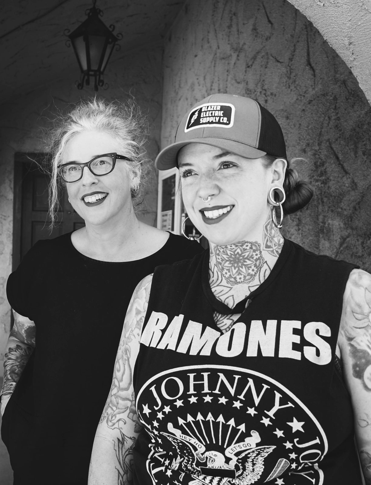 Heather Reynolds and Kate Grinstead, co-founders of Zeitgeist Tattoo, smiling side by side under a stucco archway in black and white