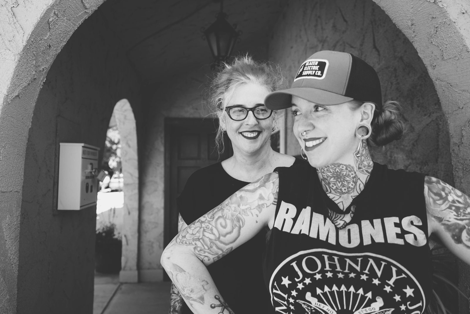 Kate Grinstead in a trucker cap and Ramones shirt laughing with Heather Reynolds beneath an archway