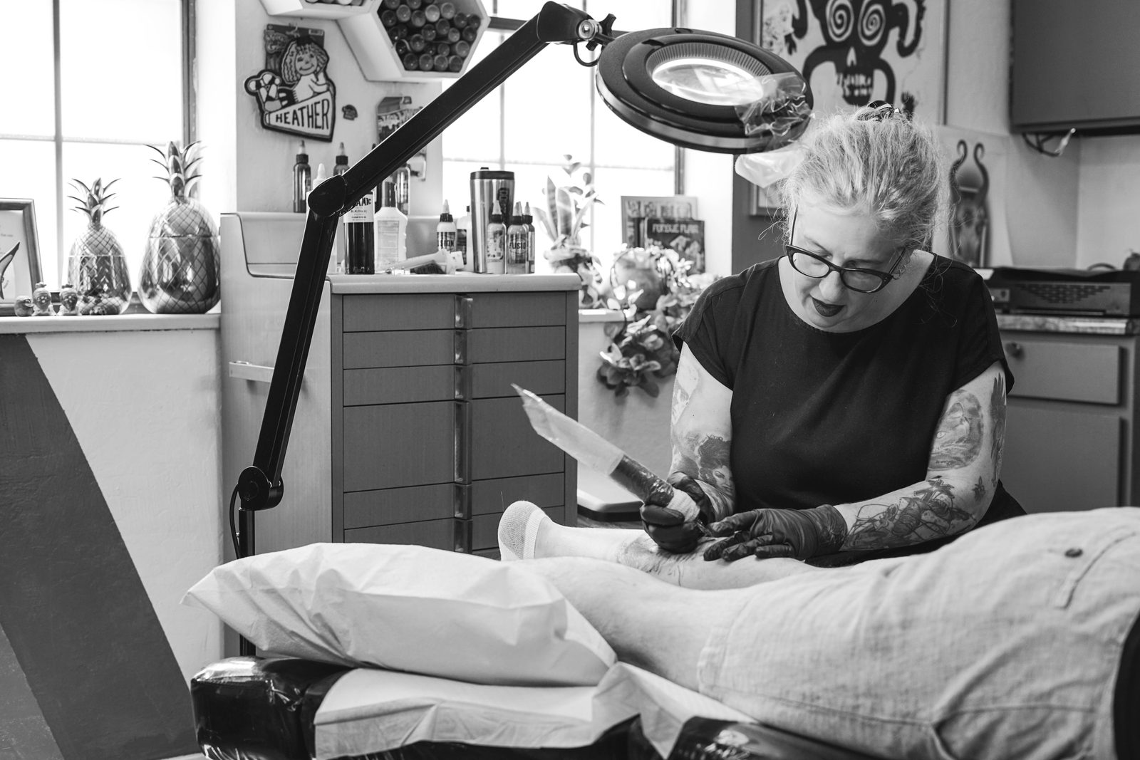 Heather Reynolds tattooing a client's leg under a magnifying lamp at her station