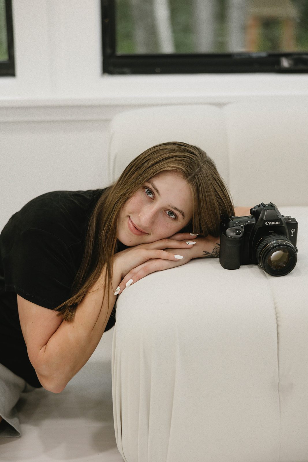 Kate Thayer, founder of Captured by Kaitlyn, resting her head on her arms on a white sofa beside her Canon film camera