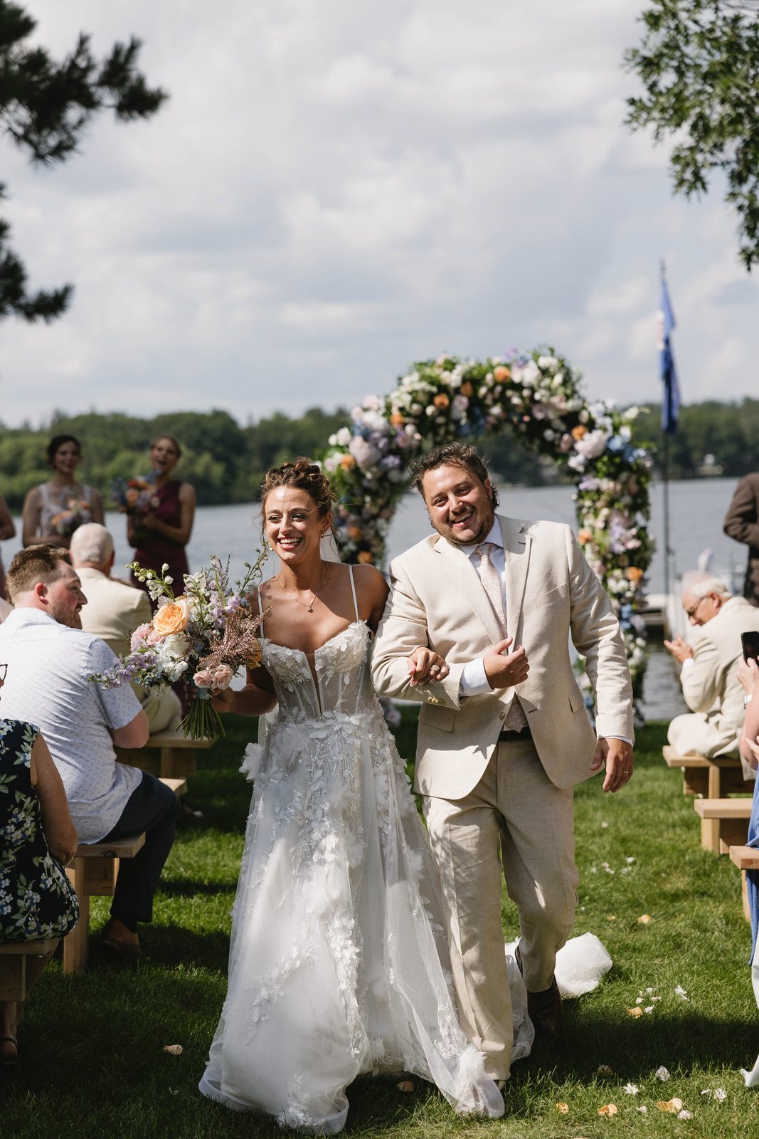 A bride and groom walking back down the aisle past their guests at a lakeside wedding, photographed by Captured by Kaitlyn