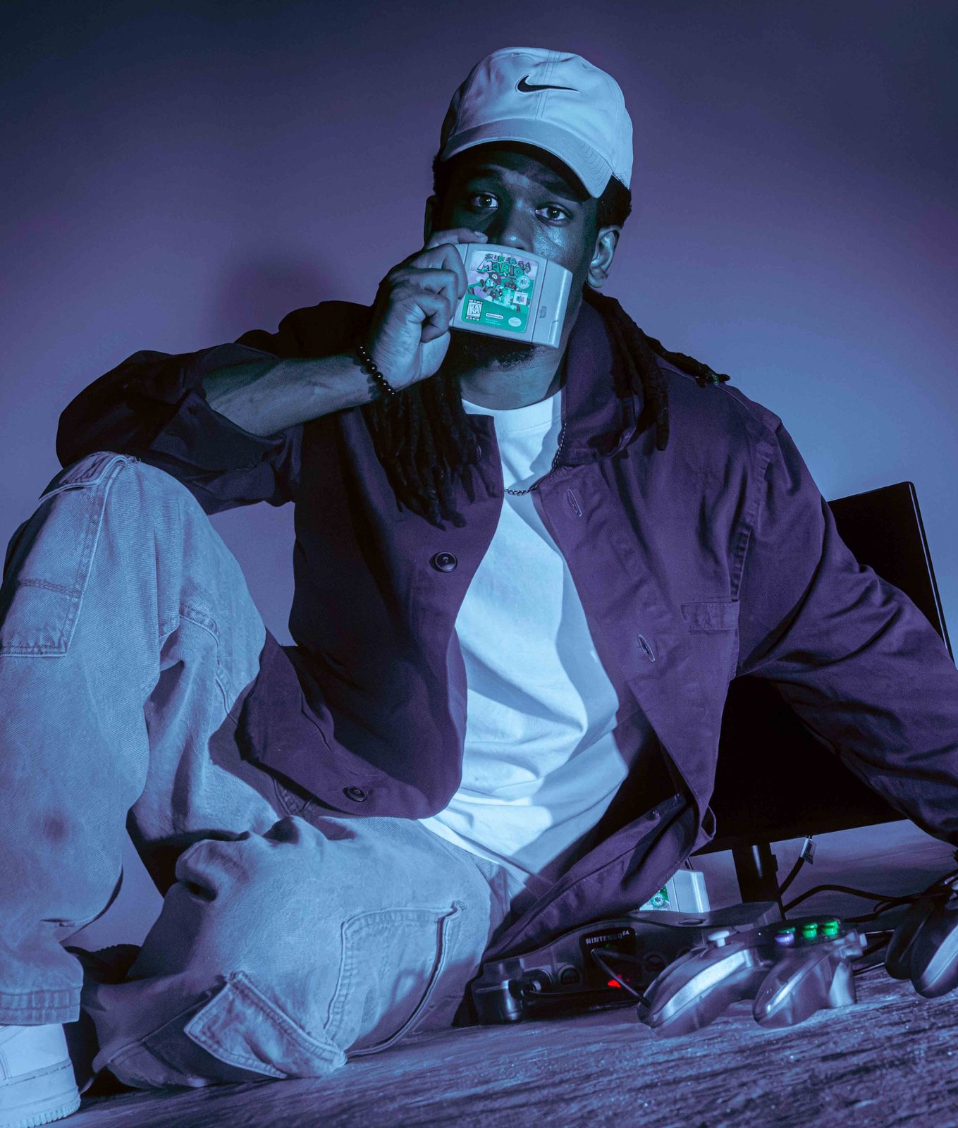 Khalid Thornton seated in a purple-lit studio holding a Nintendo 64 cartridge beside a console and controllers